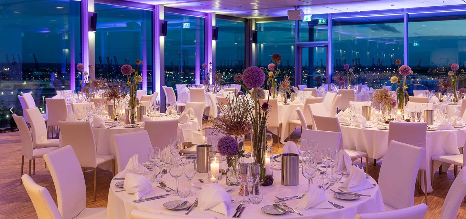 Festlich gedeckte runde Tische mit Blumendekoration vor Panoramafenstern mit Hafenblick