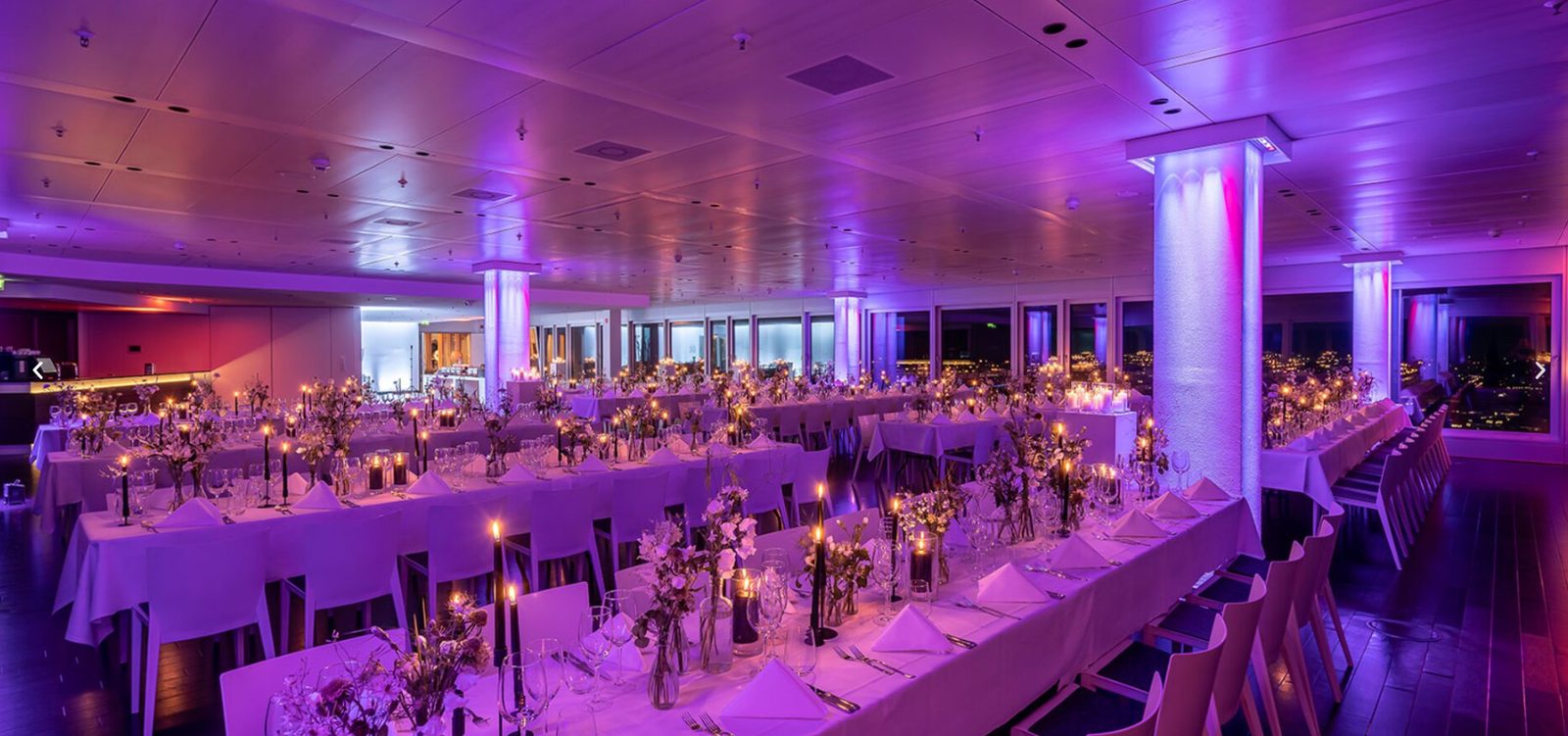 Eleganter Eventsaal von Infinity Stage mit violetter Beleuchtung, langer festlich gedeckter Tafel und Panoramafenstern